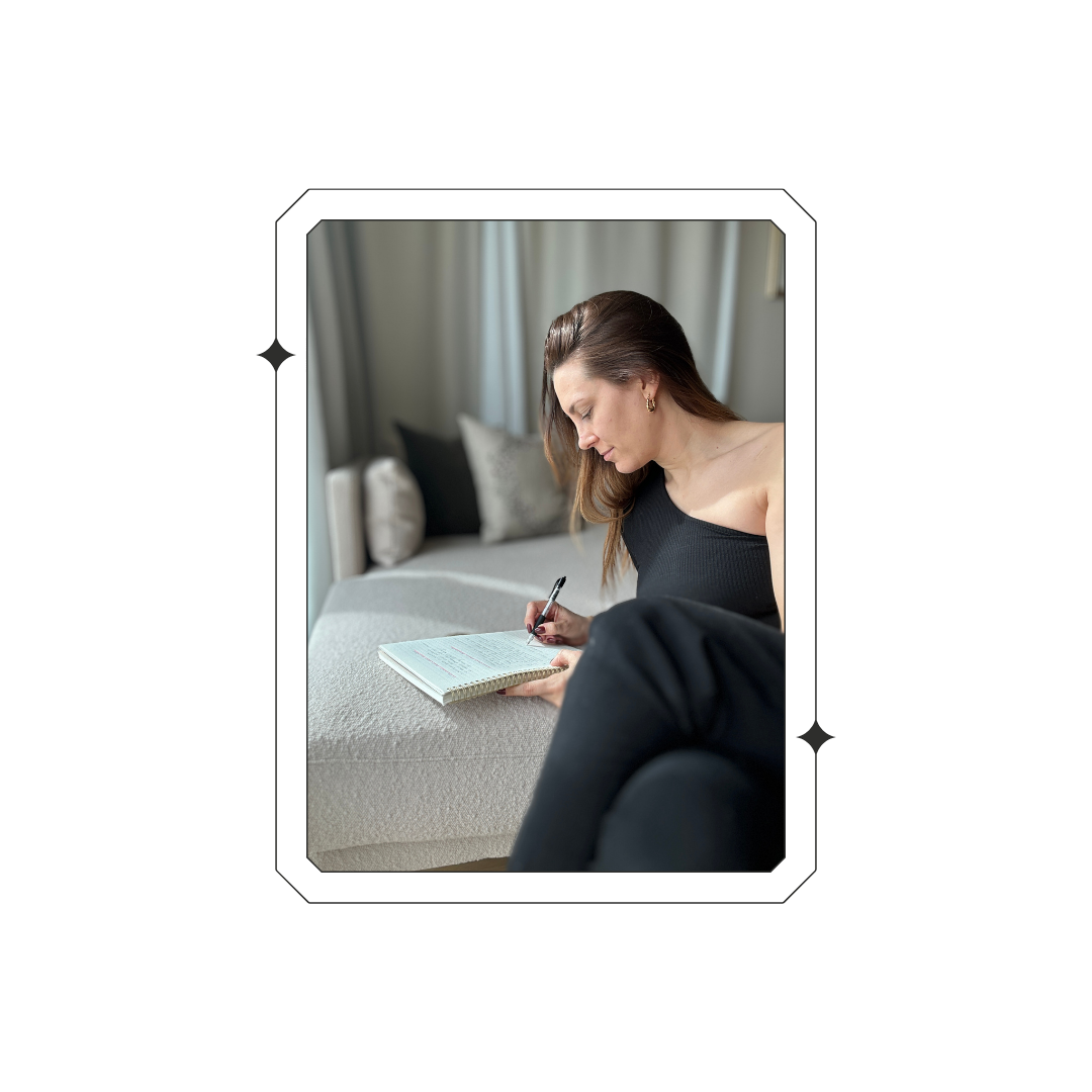 A woman writing thoughtfully in a notebook, framed on a dark background.